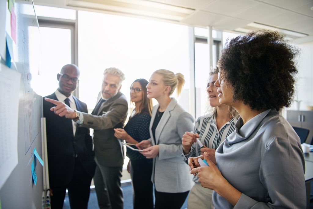 Photo of a group of business professional collaborating at a whiteboard with sunlight shining in through a window in the background.